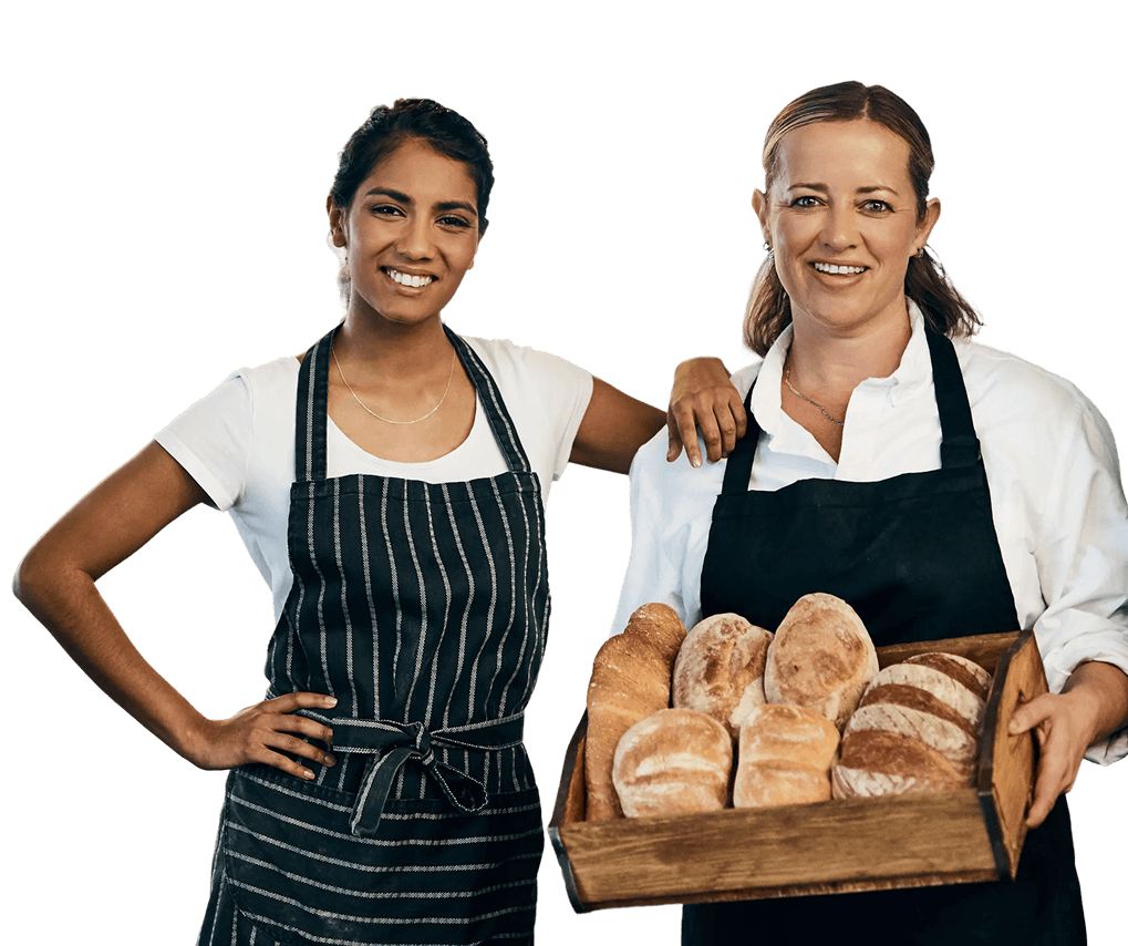Two women bakers with bread, smiling and wearing aprons, in a bakery setting.